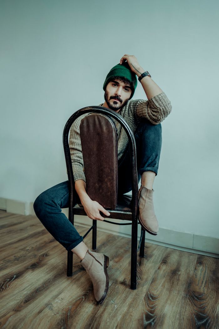 A fashionable man with a beard sits on a chair on a rustic wooden floor.