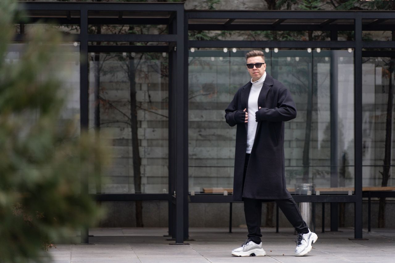 Fashionable man in sunglasses and long coat standing confidently in urban setting.